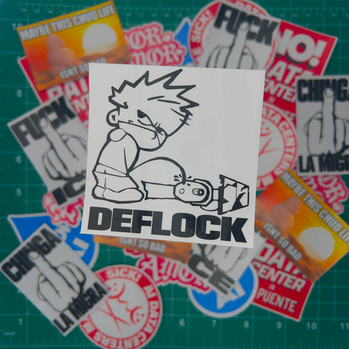 A white rectangular sticker featuring a black-and-white cartoon character sitting with a knife and the word 'DEFLOCK' in large black letters, placed on top of several other stickers on a green cutting mat.